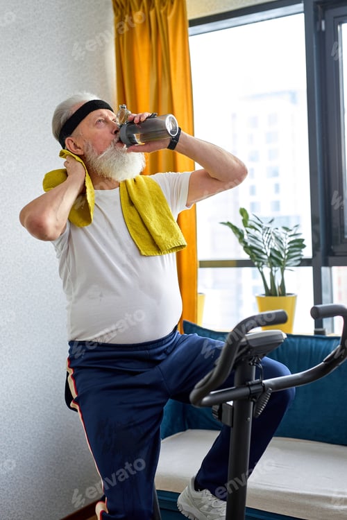 Preview: aged adult man doing physical cardio exercise on fitness bicycle, drinking water exhausted
