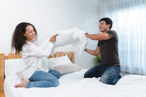 Preview: Happy Thai couple fighting by pillows on a bed and having fun together at home