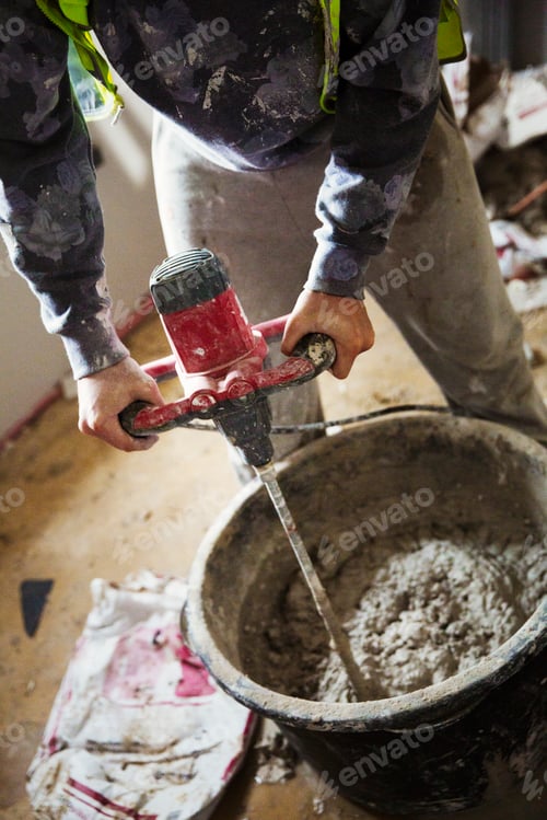 Preview: A builder mixing plaster using an electric mixer in a bucket on a construction site.