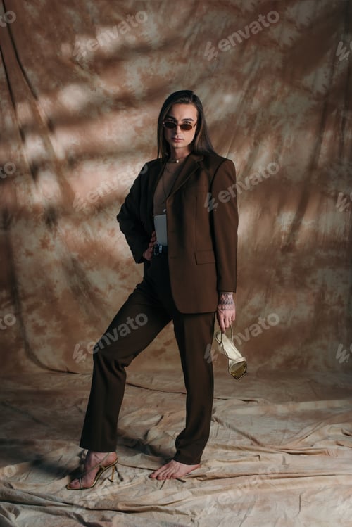 Preview: Trendy nonbinary person in suit holding shoe on abstract brown background