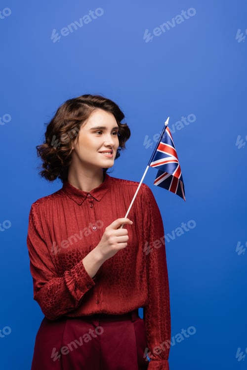 Preview: happy language teacher smiling while looking at flag of United Kingdom isolated on blue