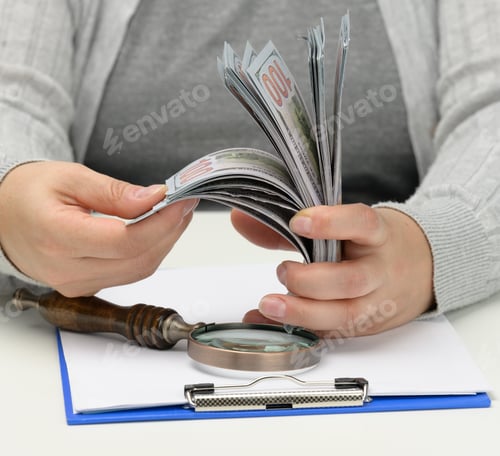 Preview: Stack of one hundred dollar bills in a woman's hand and a wooden magnifying glass
