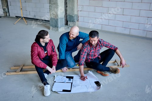 Preview: Happy young engineers sitting inside unfinished building and discussing sketch