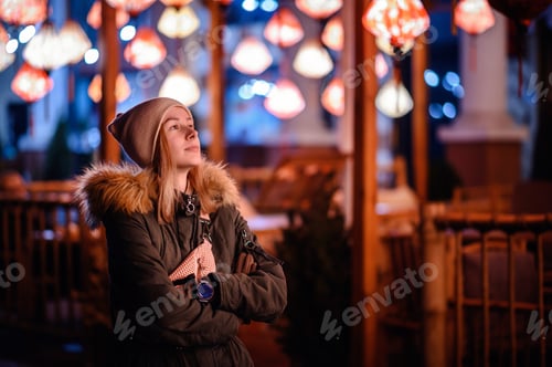 Preview: Young blonde girl in winter clothes puts on beige hat. Night street with colorful cafe lights