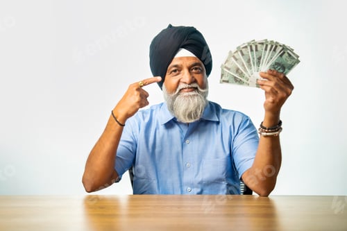 Preview: Elderly Sikh man with Indian currency fan, sitting at table, showing happy expression, success sign