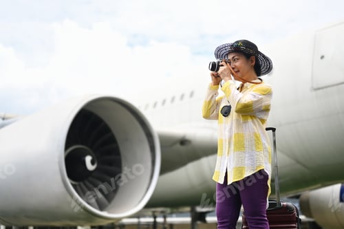Preview: Happy 60s woman in summer clothing taking photo with camera near the airplane.