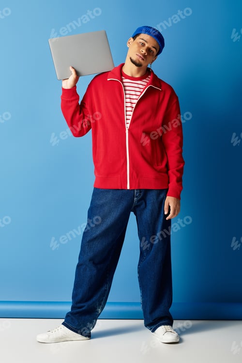 Preview: Trendy young man poses confidently with laptop against vibrant blue backdrop