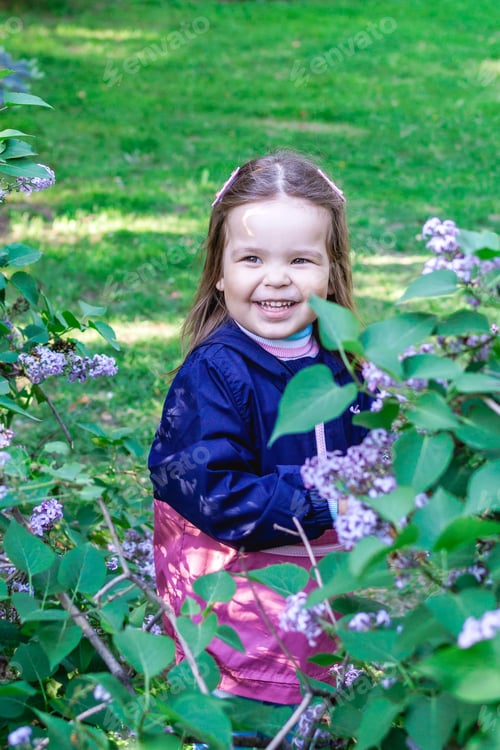 Preview: Adorable smiling little girl in a garden with lilac trees. Candid child portrait at spring season.