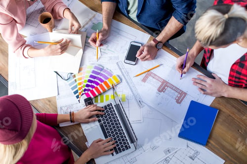 Preview: Overhead view of young male and female designers working at project with blueprints and devices