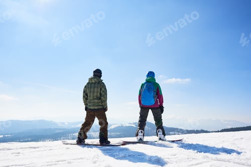 Vorschau: Rückansicht zweier Touristen-Snowboarder vor blauem Himmel und Bergen an sonnigen Wintertagen.