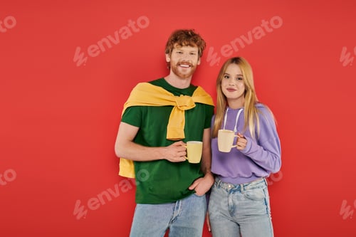 Preview: cheerful young couple holding cups of coffee on coral background, morning routine, vibrant colors,