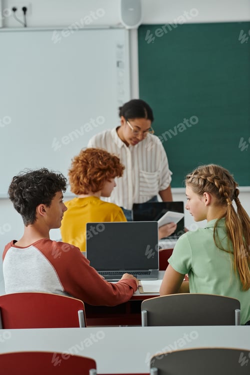 Preview: Teen classmates sitting and talking near laptop with blank screen during lesson in class in school