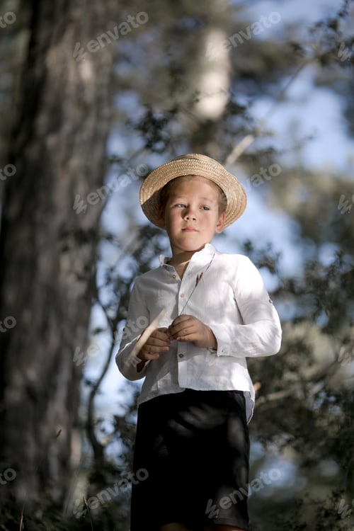 Preview: Small farm boy, smiling and looking at the camera, stands in forest. Cute kid boy in a straw hat