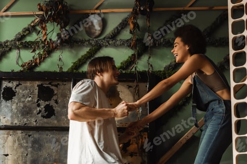Preview: side view of cheerful multiethnic couple holding hands near wall decorated with plants