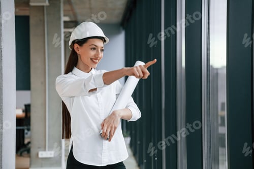 Preview: Pointing by finger to the side. Female architect in hard hat and in white clothes is indoors