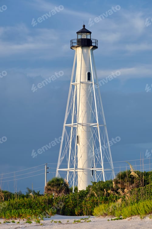 Preview: White tall lighthouse on sea shore against blue sky for commercial vessels navigation