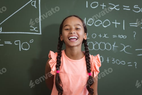 Preview: Portrait of laughing mixed race schoolgirl standing in front of chalkboard in classroom maths lesson
