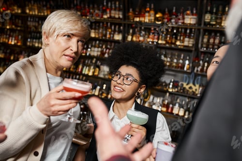 Preview: middle aged woman with cocktail glass talking to multicultural colleagues spending time in bar