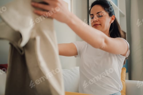 Preview: Woman holding beige clothes at home decluttering her wardrobe