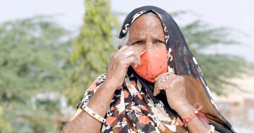 Preview: Closeup of an elderly Indian woman fixing her vibrant orange Covid mask while wearing a headscarf