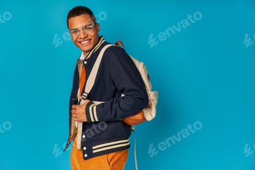 Preview: carefree african american student in jacket and orange pant, with backpack looking at camera on blue