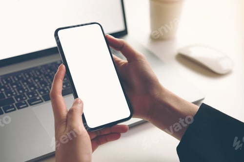 Preview: Woman holding smartphone mockup of blank screen and laptop on the table.