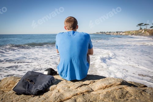 Preview: Young man sitting by rock, looking out to sea, rear view