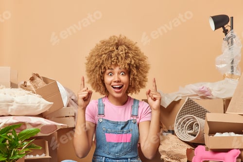 Preview: Surprised curly haired woman dressed casually indicates index fingers overhead dressed in casual t