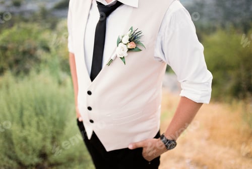Preview: Groom in a vest with a boutonniere stands with his hands in his pockets. Cropped