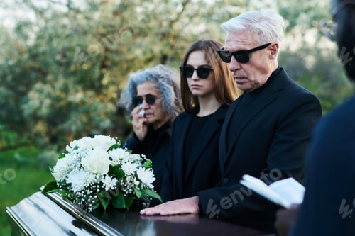 Preview: Mature man in sunglasses and black attire keeping hand on lid of closed coffin