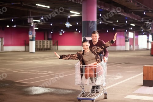 Preview: ↟ ✓ Cheerful happy young couple in casual clothes having fun and riding on a shopping cart