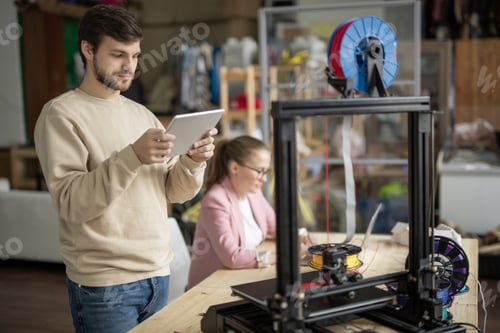 Preview: Young man looking at online figure on touchpad display in front of 3d printer