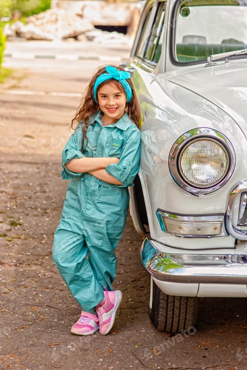 Preview: A cute little girl in a blue jumpsuit stands next to a retro car in the summer on a sunny day