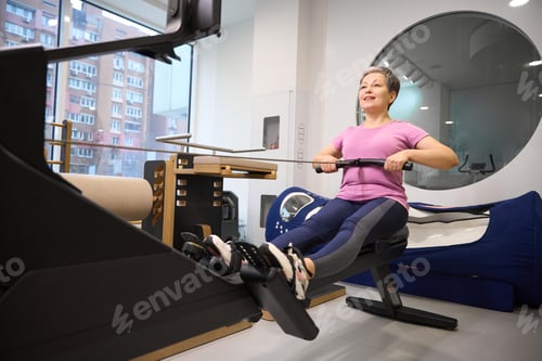 Preview: Mature smiling caucasian woman doing sport exercise for back on rowing trainer