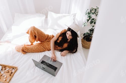 Preview: Beautiful young stylish woman in white hat lying on bed and using laptop computer