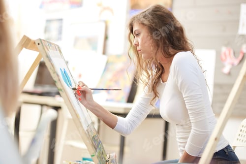 Preview: Young pretty girl with brown curly hair dressed in white blouse paints a picture at the easel in the