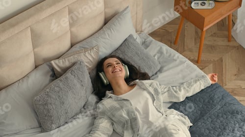 Preview: Top view young happy brunette woman lying on bed headphones listening music using smartphone mobile
