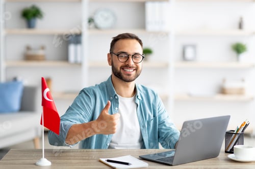 Preview: Happy young guy sitting at desk with flag of Turkey, using laptop, showing thumb up gesture at home