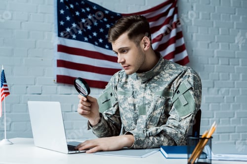 Preview: young soldier in uniform holding magnifying glass near laptop in office