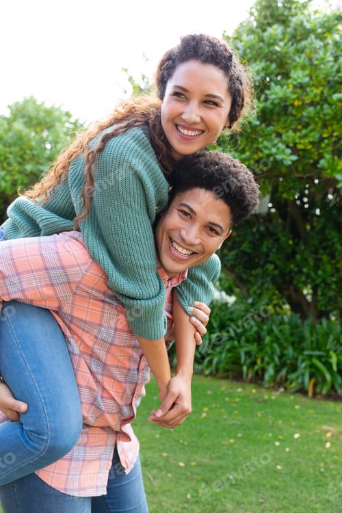 Preview: Portrait of happy biracial couple having fun piggybacking in garden