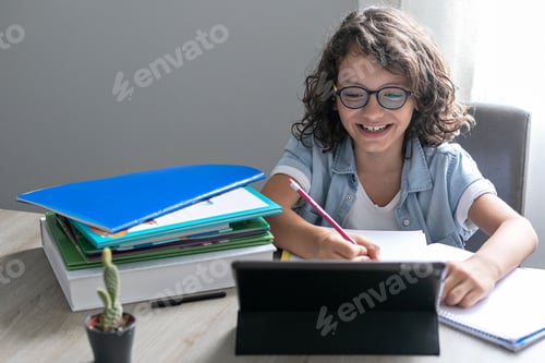 Preview: Little adorable school boy with eyeglasses, studying online from home. kid using digital tablet