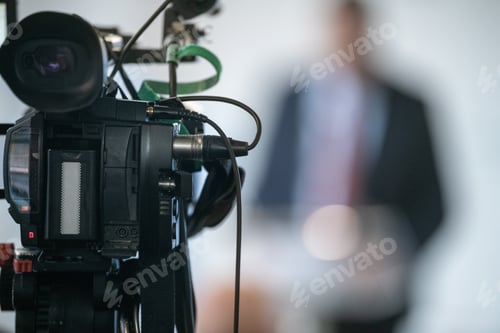 Preview: TV camera capturing live press conference with officials. Emphasizes clarity