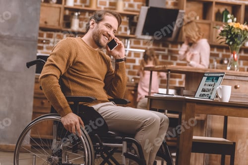 Preview: cheerful disabled man in wheelchair talking on smartphone and using laptop at home