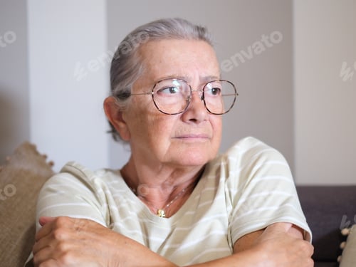 Preview: Portrait of pensive senior woman with gray hair and eyeglasses sitting on home sofa