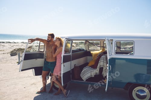 Preview: Front view of young Caucasian couple standing near camper van at beach in the sunshine