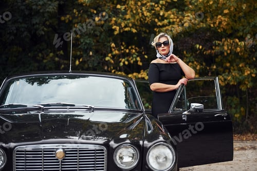 Preview: Blonde woman in sunglasses and in black dress near old vintage classic car