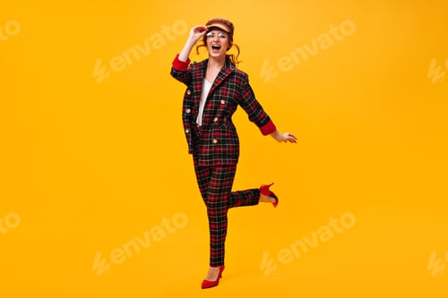 Preview: Pretty girl in eyeglasses, cap and suit jumps on orange background. Joyful woman in cap and round e