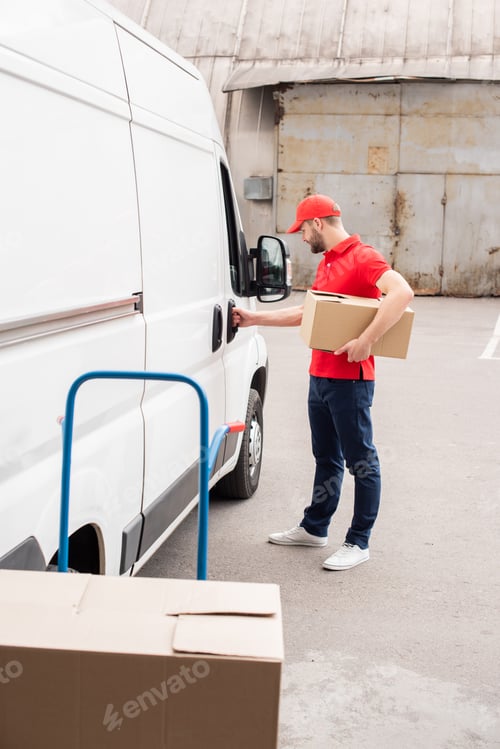 Preview: side view of young delivery man in uniform with cardboard box standing near white van in street