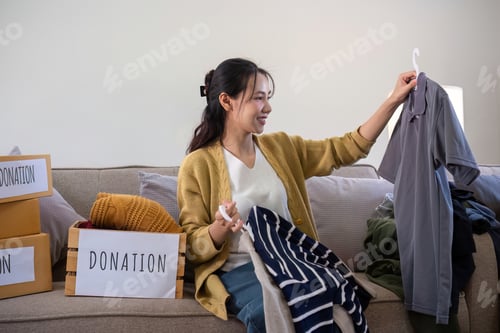 Preview: Cute Asian woman sits on a sofa sorting unwanted clothes to donate.