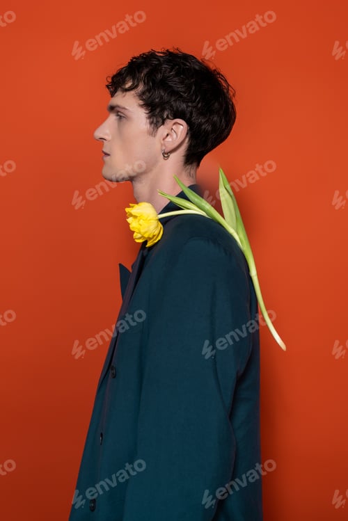 Preview: Side view of trendy brunette man with yellow tulip on shoulder on red background